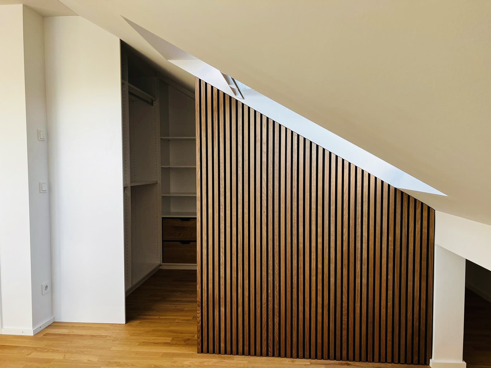 Gesamtansicht des begehbaren Kleiderschranks unter der Dachschräge: Raumteiler am Fenster, klassischer Kleiderschrank mit Kleiderstangen an der (fast) senkrechten Wand und Regal mit Schubladen an der Rückwand mit Dachschräge. Realisiert von unserem Münchener Team.